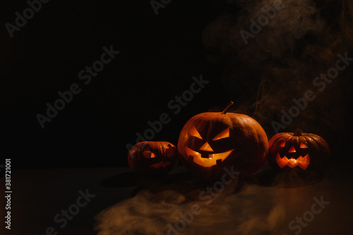 Three creepy halloween steaming pumpkins with a carved luminous smirk on a black background. A handmade jack-o-lantern head with a candle inside in the dark among the fog. Trick or treat. Postcard.
