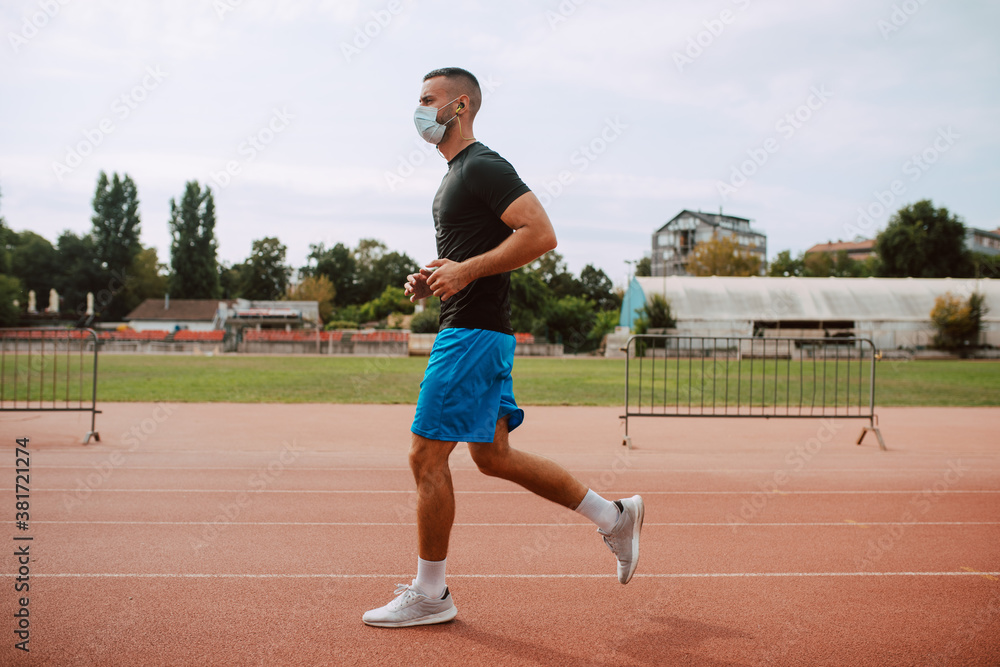 A shot of a young male caucasian athlete  with face mask training on a racetrack. The sprinter runs on athletic all-weather running track. COVID - 19 coronavirus protection