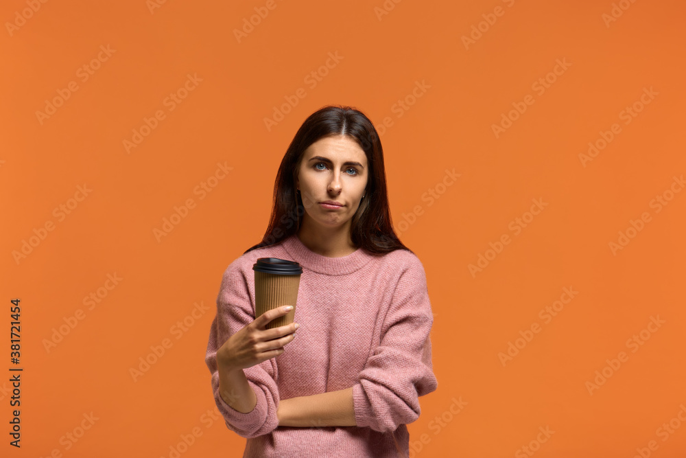 Need to cheer up. Tired sleepy beautiful woman in pink sweater with a tortured look has sad expression, carries disposable cup of drink containing caffeine. on orange background Need to cheer up. Tired sleepy beautiful woman in pink sweater with a tortured look has sad expression, carries disposable cup of drink containing caffeine. on orange background