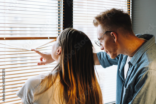 Fotografie Young cheerful couple looks out the window through the blinds, looks at what is happening outdoor