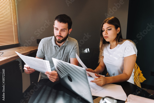 A young couple manages finances by looking through their bank accounts in the living room at desk using laptop. Woman and man look at documents together. Planning budget expenses.