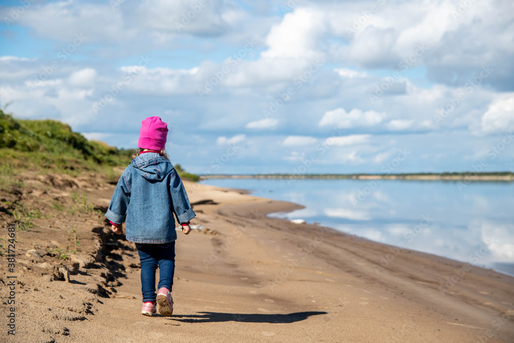 The child is alone on the sandy bank of the river. A girl in jeans, a denim jacket and a pink hat walks along the sandy bank of the river. View from the back. Child safety concept.