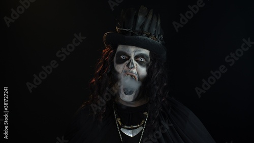 Sinister man with Halloween skeleton makeup exhaling cigarette smoking from his mouth and smiling