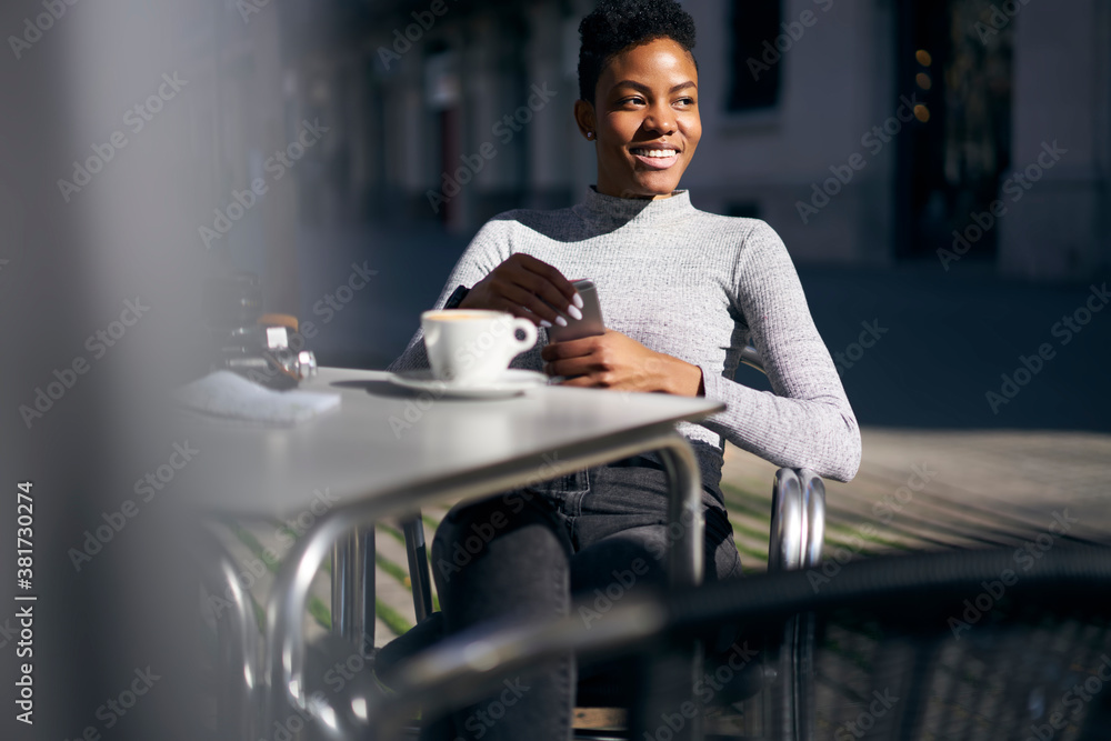 Fototapeta premium Crop cheerful young black woman browsing smartphone at table in street cafeteria