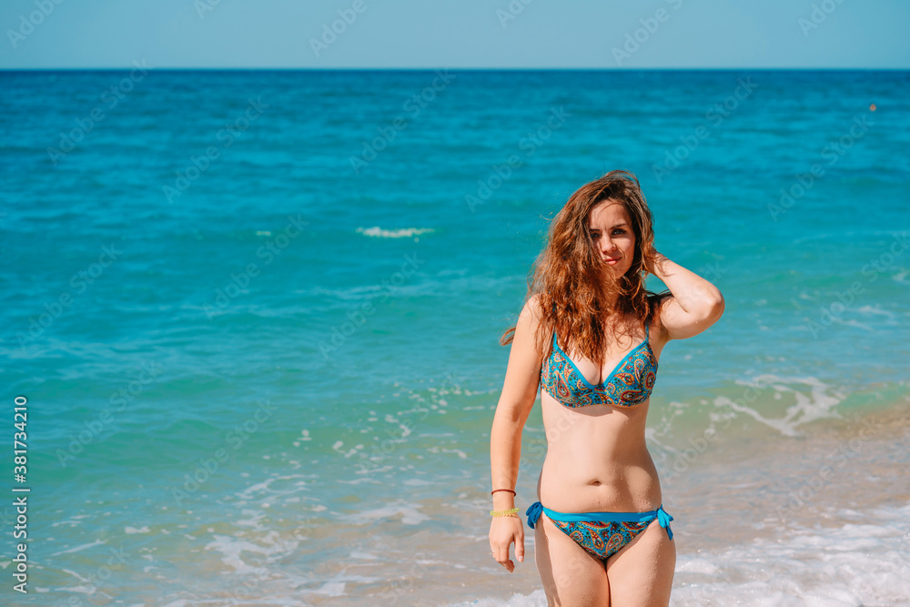 A young woman in a bright swimsuit and long hair walks on the sand along the sea with waves, clear azure water