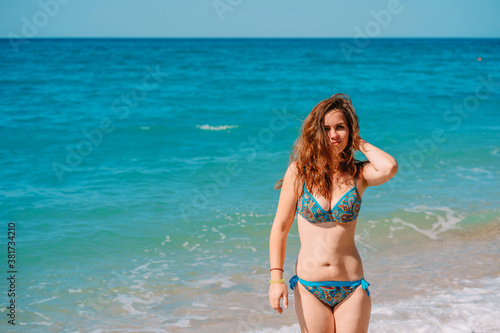 A young woman in a bright swimsuit and long hair walks on the sand along the sea with waves, clear azure water