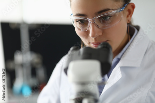 Chemist in goggles and white coat looks through microscope. Conducting scientific laboratory experiments concept.