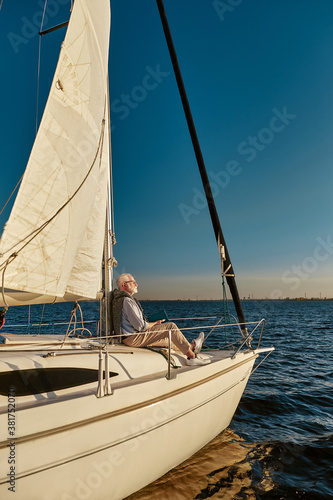 Vertical shot of carefree retired man enjoying amazing view and relaxing while sitting on the side of his sailboat or yacht deck floating in the calm blue sea © Kostiantyn