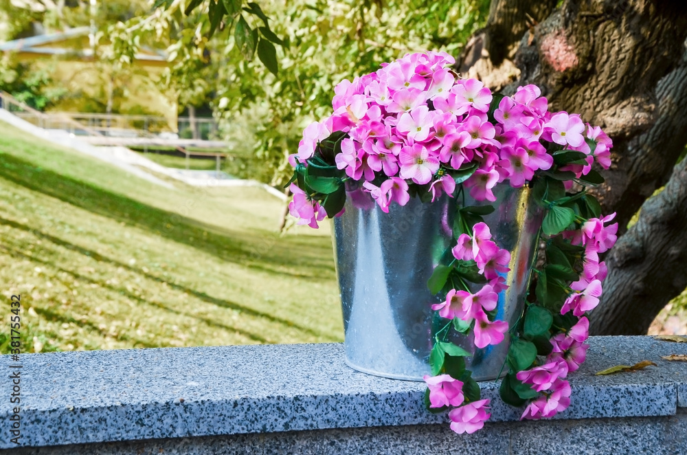 Metal bucket with pink flowers on a granite parapet against the backdrop of trees and a park. Outdoor cafe decor element. Space for text. Selective focus and blurred background