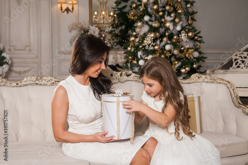 Merry Christmas and Happy Holidays. Cheerful mom and her cute daughter girl exchanging gifts in white classic interior against the background of a piano and a decorated Christmas tree. New Year 2021.