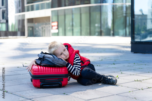 Tired little boy traveler on the background of modern buildings lies on a suitcase