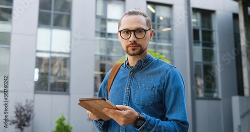Portrait of Caucasian young man in glasses tapping on tablet device and smiling to camera at city street. Handsome male texting message and browsing on gadget computer outdoors. Gadget user concept.