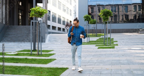Caucasian young man walking steps downn at street, tapping on smartphone and sipping drink. Male gooing down the ladder outdoor, texting mesage on phone and drinking coffee to-go. Rushing in morning. photo