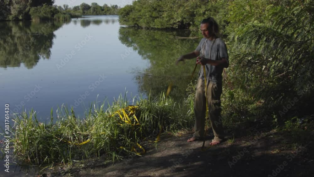 Caucasian man sets slackline between trees above the river. A man practices highlining over water in summer at dawn. Caucasian man sets slackline between trees above the river. A man practices highlining over water in summer at dawn.