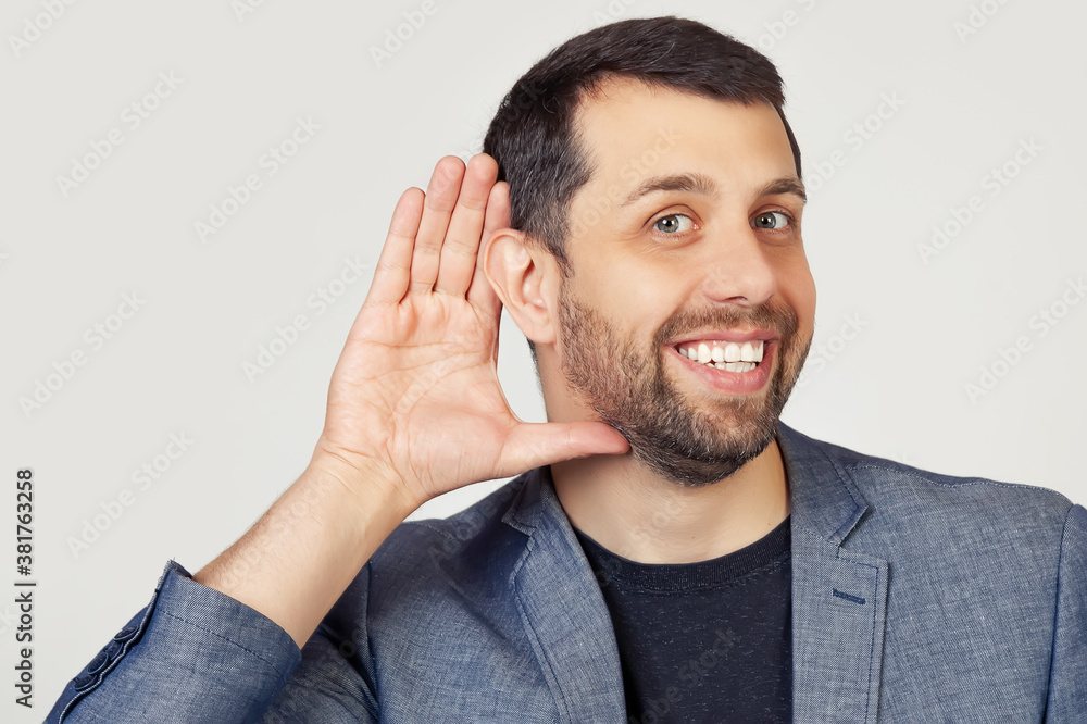 Young businessman man with a beard in a jacket, smiling with his hand above his ear, listening to rumors or gossip. Deafness concept. Portrait of a man on a gray background Young businessman man with a beard in a jacket, smiling with his hand above his ear, listening to rumors or gossip. Deafness concept. Portrait of a man on a gray background