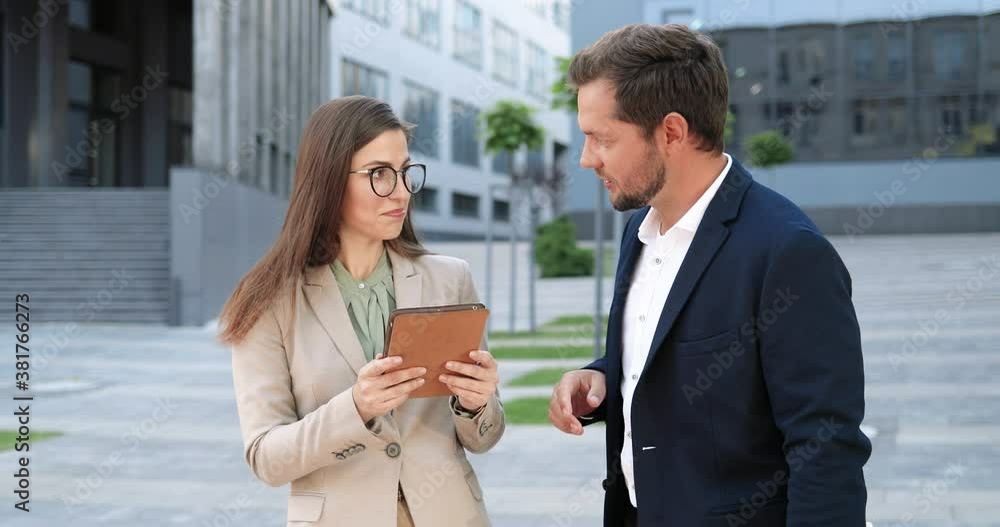 Caucasian male and female colleagues standing in city and discussing some document on tablet device. Business partners talking and using gadget outdoor. Man and woman having discussion in town. Caucasian male and female colleagues standing in city and discussing some document on tablet device. Business partners talking and using gadget outdoor. Man and woman having discussion in town.