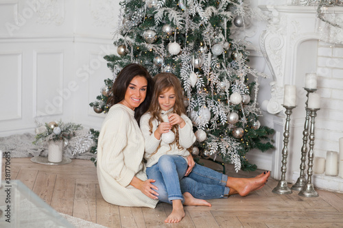Happy family on New Year Eve. Young beautiful mother and little cute daughter in white cozy sweaters and blanket have fun and hug under a decorated Christmas tree. Festive home classic interior © Алина Троева