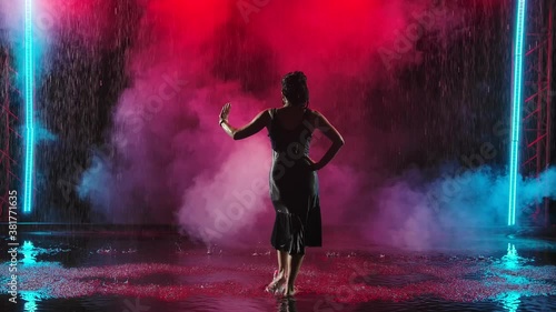 A passionate performance of Spanish flamenco dance in the rain. A charming young woman dances on the surface of the water and creates many splashes. Silhouette. Slow motion.