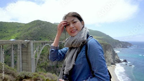 woman travel photographer blogger with camera sightseeing bixby bridge highway 1 standing on mountains in big sur. young girl turn around smiling face camera while visiting nature on summer trip usa.