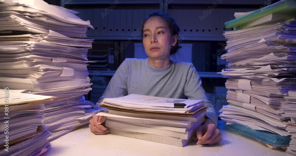 Exhausted asian business woman putting documents on desk cover with stack of paperwork. Tired alone girl looking at paper and lay down on pile of sheets while working hard at late night at home.