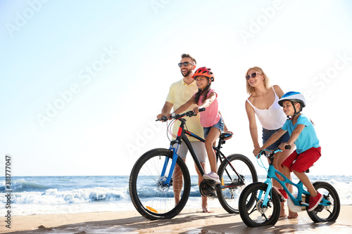 Happy parents teaching children to ride bicycles on sandy beach near sea