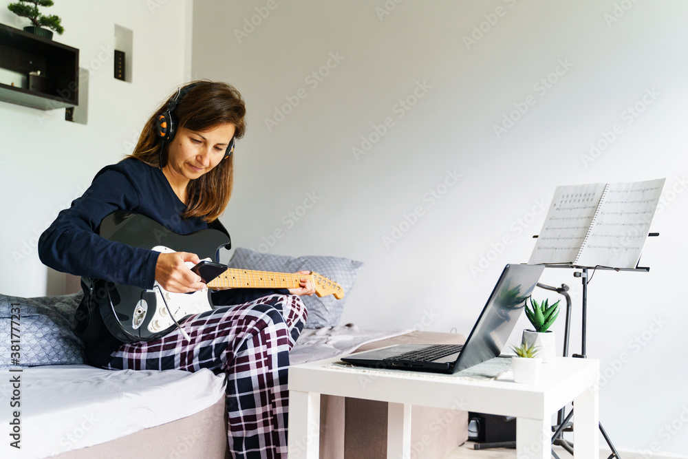 Adult caucasian woman sitting on bed with earphones on head using mobile phone to play music while holding guitar in her bedroom at home - real people leisure weekend activities concept copy space