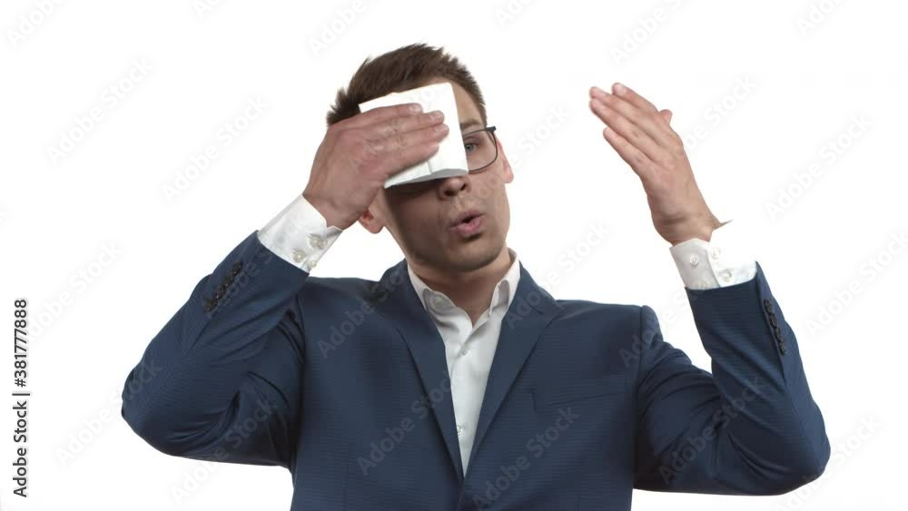 Handsome young businessman with blond hair, wearing blue suit and glasses, feeling hot, sweating and wiping sweat off forehead with napkin, standing over white background Handsome young businessman with blond hair, wearing blue suit and glasses, feeling hot, sweating and wiping sweat off forehead with napkin, standing over white background