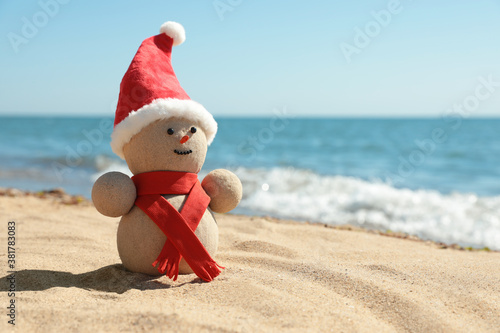 Snowman made of sand with Santa hat and scarf on beach near sea, space for text. Christmas vacation