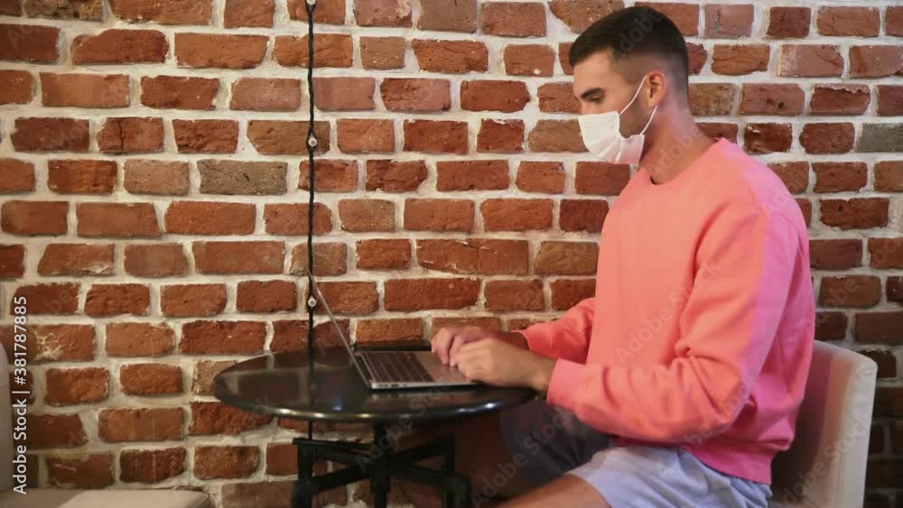 Young businessman in medical mask is sitting in a cafe and working on a laptop Young businessman in medical mask is sitting in a cafe and working on a laptop