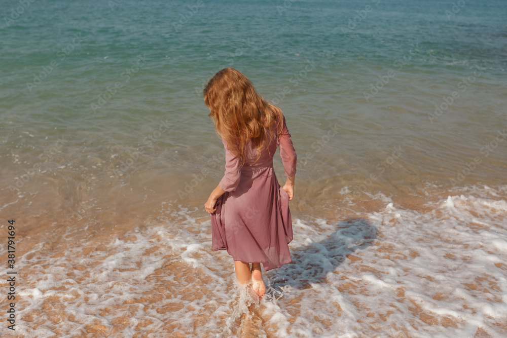 Back view  trendy girl hold her long summer dress trying not to wet it in waves. Summer vacation by the sea