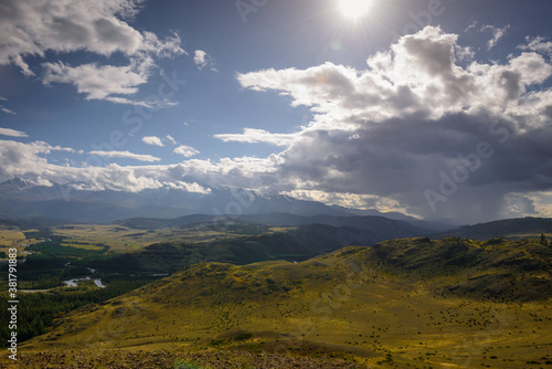 Majestic panorama of mountain plain on the background of snow-covered ridge before thunderstorm. Sun's rays break through huge clouds and beautifully illuminate green steppe and winding river.