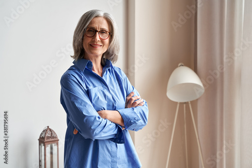 Smiling confident mature older woman standing arms crossed indoors looking at camera. Stylish classy middle aged senior 60s grey-haired lady wearing blue elegant shirt and glasses indoors, portrait. photo