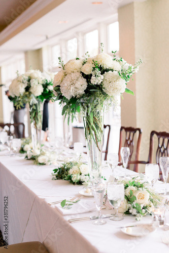 A pretty table decorated with two tall vases with a white and green flower arrangement centerpiece. 