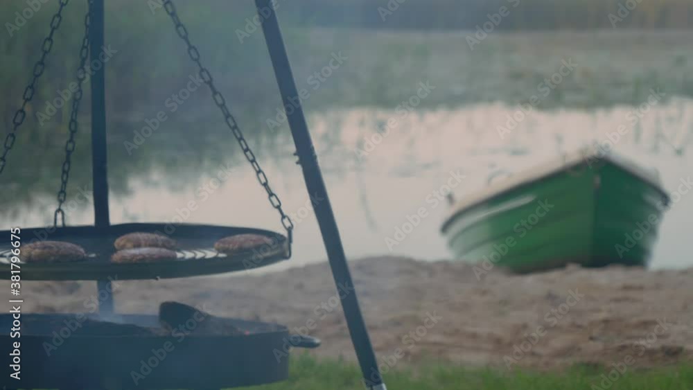 Grilling Burger Beef Patties On A BBQ Tripod Swivel Hanging Grill Near The Lake With Wooden Boat On Shore. - wide shot