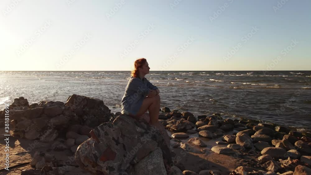 Aerial orbiting: Attractive woman sits on rocks at Cape Kolka beach looking at beautiful sea Aerial orbiting: Attractive woman sits on rocks at Cape Kolka beach looking at beautiful sea