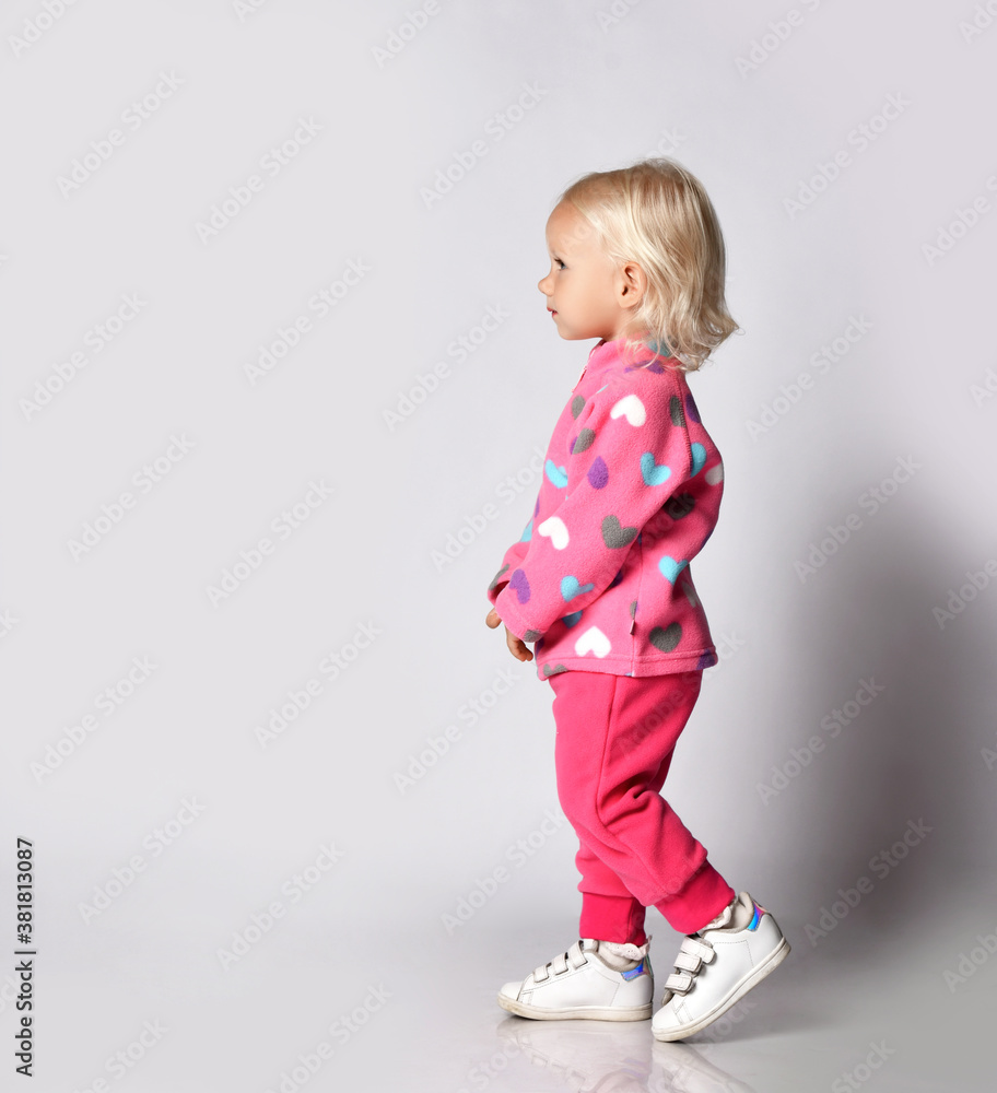 Blonde baby kid girl in pink warm fleece clothing with heart print pattern walks passes by camera. Side view Blonde baby kid girl in pink warm fleece clothing with heart print pattern walks passes by camera. Side view