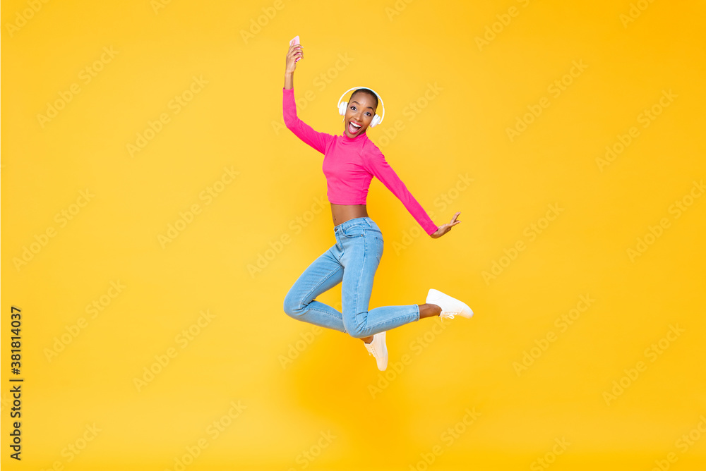 Fun portrait of smiling energetic African American woman wearing headphones listining to music from smartphone and jumping isolated on colorful yellow background