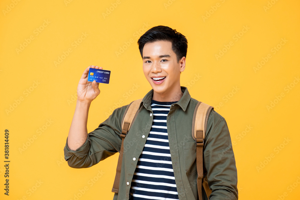 Waist up portrait of smiling young Asian backpacker tourist man showing credit card in isolated studio yellow background Waist up portrait of smiling young Asian backpacker tourist man showing credit card in isolated studio yellow background