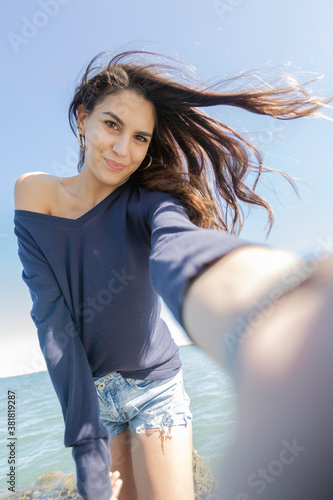 Vertical shot selfie of young brunette italian woman long hair dark blue sweater romantic hair in the wind happy, smiling and with the intense blue sky and with a flower in her right hand holding down