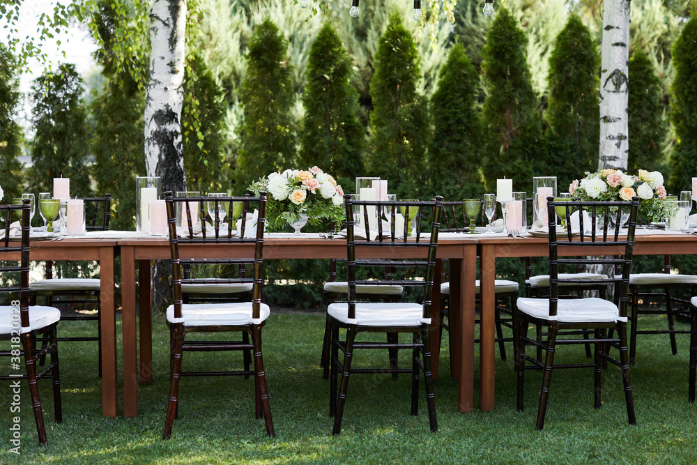 chairs and dinner tables with white cloth, served with porcelain and green glasses. Georgeous wedding or event party table decorated with flowers on a green lawn