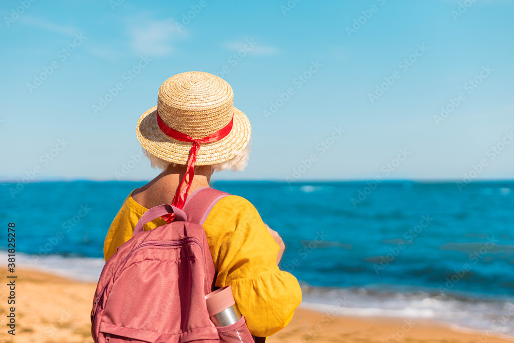 Stylish woman traveler in a summer straw hat with a tourist backpack and a thermo mug back stands and looks at the beautiful seascape