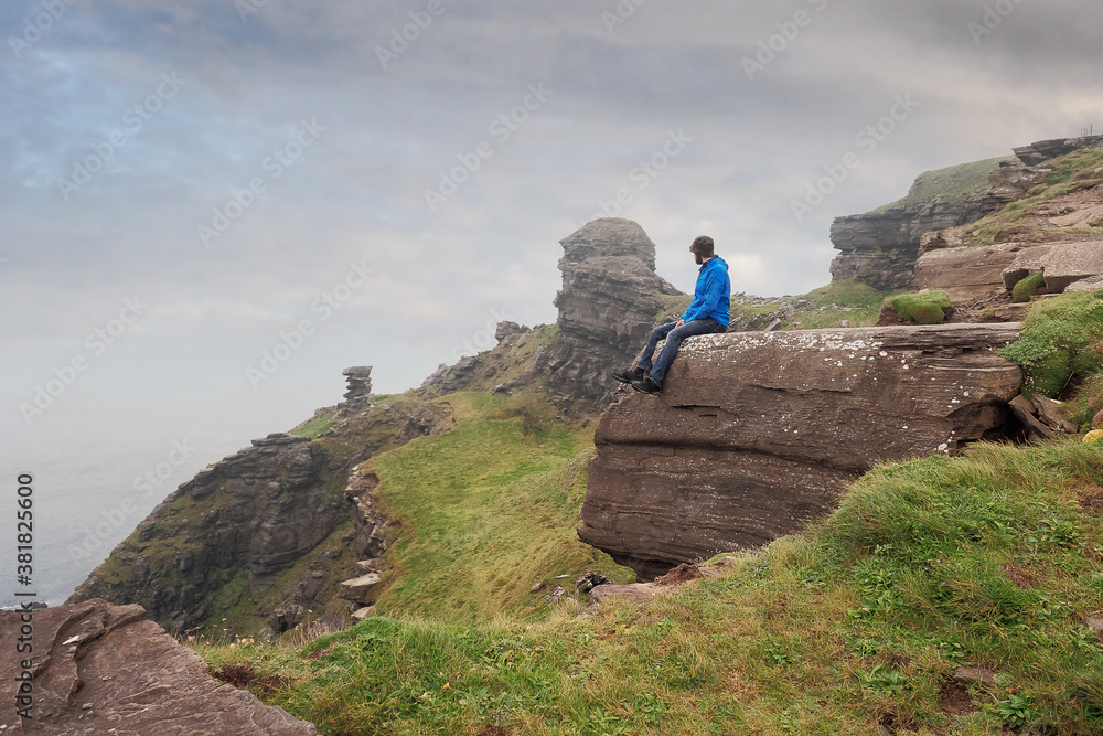 Man sitting on a edge of a cliff and looking at beautiful and rough terrain around him. Cliffs of Moher, county Clare, Ireland. Cloudy sky. Concept taking risk on vacation