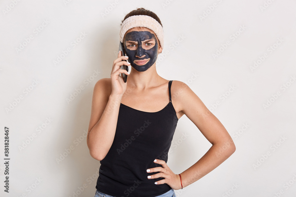 Young teen girl with black clay facial mask making phone call, standing isolated over white background, wearing headband and t shirt, keeping hands on hip, looks at camera.