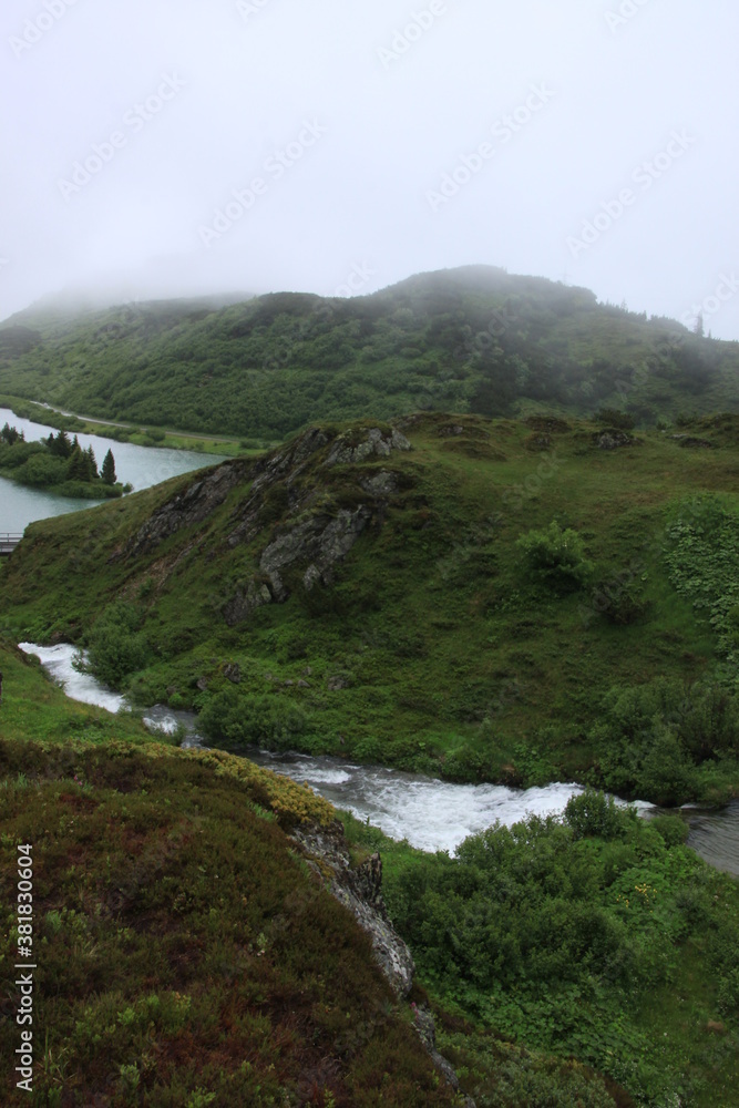 Berglandschaft mit Wiese und grüner Alm und See und Fluss im Nebel mit Wolken