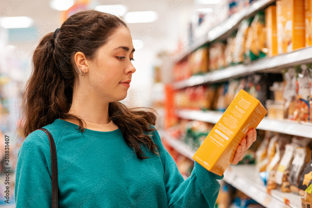 Portrait of a young Caucasian beautiful woman who reads the ingredients on the product packaging. Shelves with goods in a blur in the background. The concept of buying goods and shopping - obrazy, fototapety, plakaty 