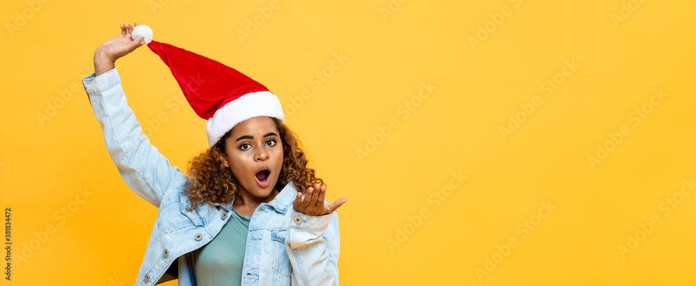 Surprised African American woman wearing Christmas hat with open palm gesture on yellow isolated banner background with copy space Surprised African American woman wearing Christmas hat with open palm gesture on yellow isolated banner background with copy space