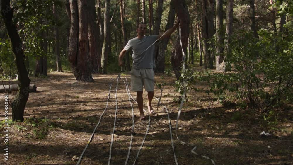 Caucasian man sets slackline between trees above the river. A man practices highlining over water in summer at dawn. Caucasian man sets slackline between trees above the river. A man practices highlining over water in summer at dawn.