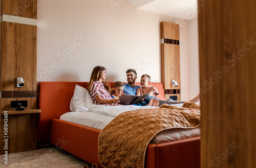 Fotografie Family of four enjoying together at hotel room, sitting at bed reading hotels offers