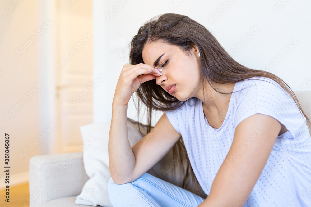Portrait of an attractive woman sitting on a sofa at home with a headache, feeling pain and with an expression of being unwell. Upset woman sitting on couch feeling strong headache migraine. Portrait of an attractive woman sitting on a sofa at home with a headache, feeling pain and with an expression of being unwell. Upset woman sitting on couch feeling strong headache migraine.