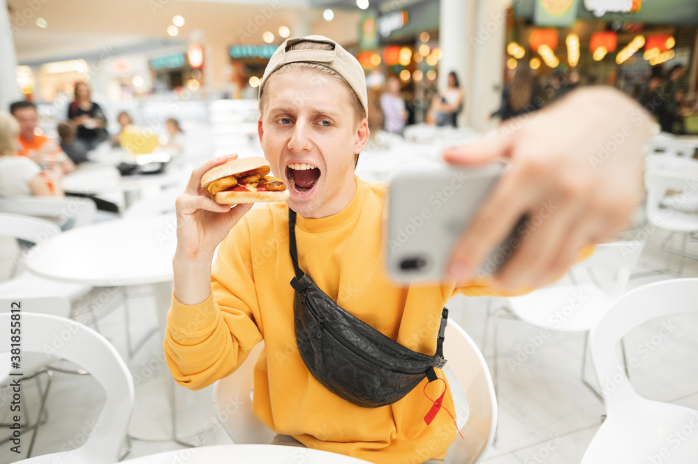 Happy young man sitting at fast food restaurant eating burger and taking selfie. A guy in casual clothes poses on a smartphone camera with a burger in his hand, takes selfies with fast food. Happy young man sitting at fast food restaurant eating burger and taking selfie. A guy in casual clothes poses on a smartphone camera with a burger in his hand, takes selfies with fast food.